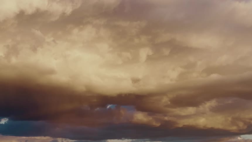 A closeup view shows heavy voluminous storm clouds in shades of brown and orange moving slowly across the sky with small patches of blue visible between them