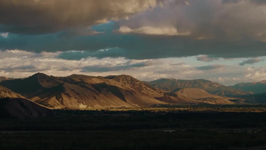 A wide landscape view shows rolling brown mountains and dark valleys partially illuminated by a patch of late afternoon sunlight shining through dramatic cloudy sky