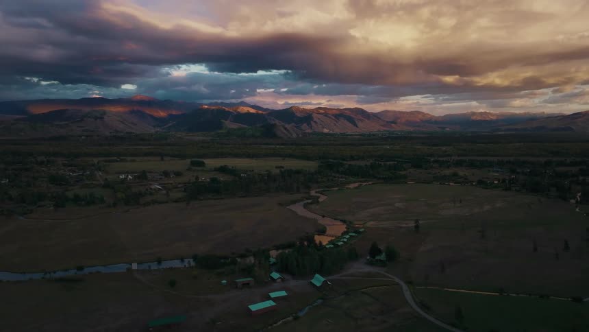 A dynamic highangle view that slowly rises and moves forward over a valley containing fields a winding waterway and a small group of structures all backed by a range of mountains under a sunset sky with heavy clouds