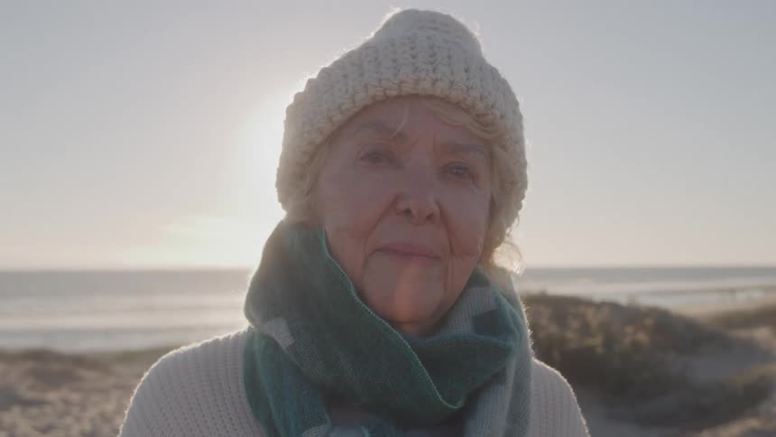 Elderly woman by the sea at sunset wearing knit hat and blue scarf