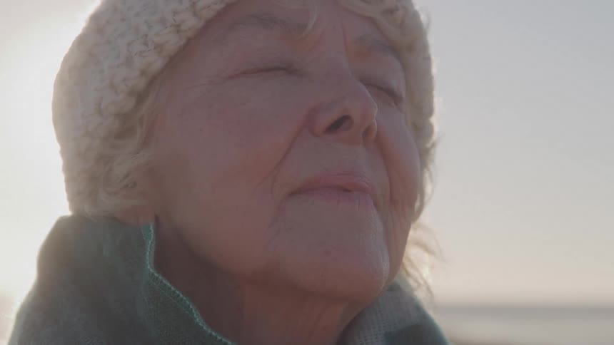 A close view shows an older womans face tilted up with her eyes closed as she wears a knit hat and a blanket and enjoys the warm sunlight