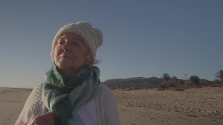 A medium close view shows an older woman wearing a white knit cap white cardigan and green scarf standing on a sandy beach with mountains and palm trees in the background under a clear sky