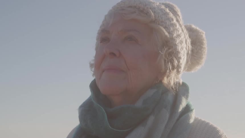 Elderly woman wearing a hat and scarf looks upward on a clear day outdoors
