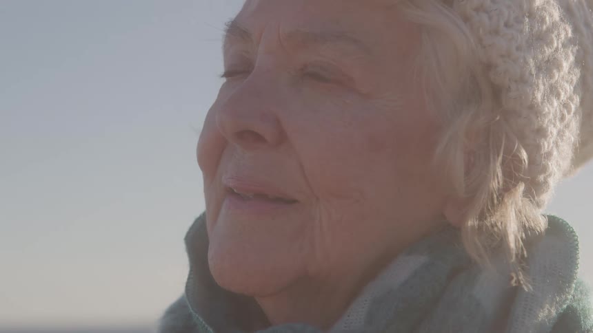An elderly woman in a knit hat and scarf gazes upward in warm sunlight looking calm and hopeful