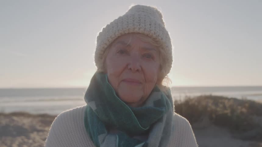 Elderly woman wearing a hat and scarf smiles softly by the seaside on a sunny day