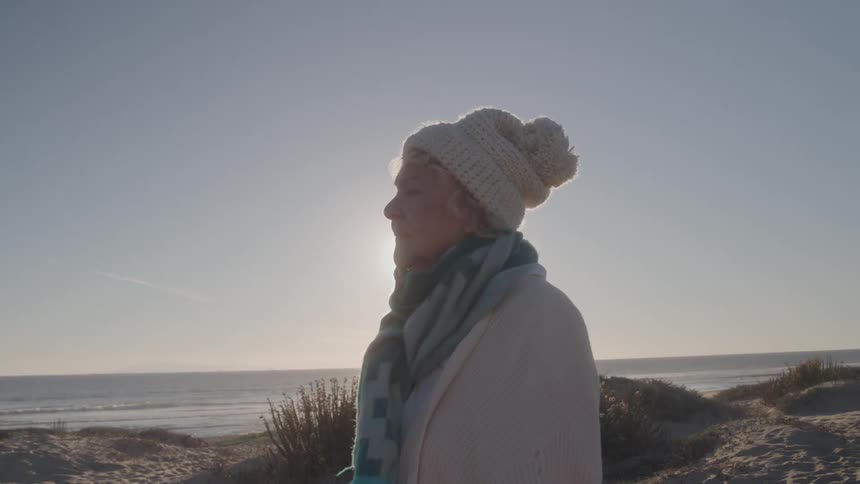 A medium side view shows an older woman wearing a white knit hat and a shawl standing on a sandy dune looking out at the ocean with the bright sun creating a strong backlight