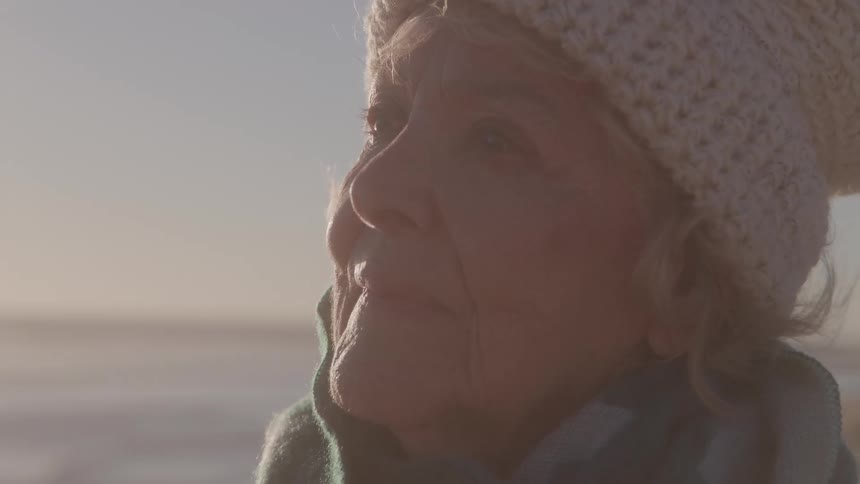 A closeup side view shows an older woman wearing a white knit cap and a teal shawl smiling with her eyes open as she looks towards the ocean with warm backlighting