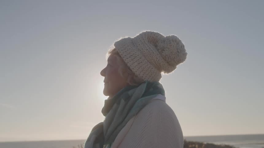 Elderly woman wearing a hat and scarf gazes at the horizon on a sunny day