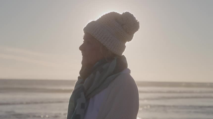 Elderly woman wearing a hat and scarf smiles while enjoying the seaside view