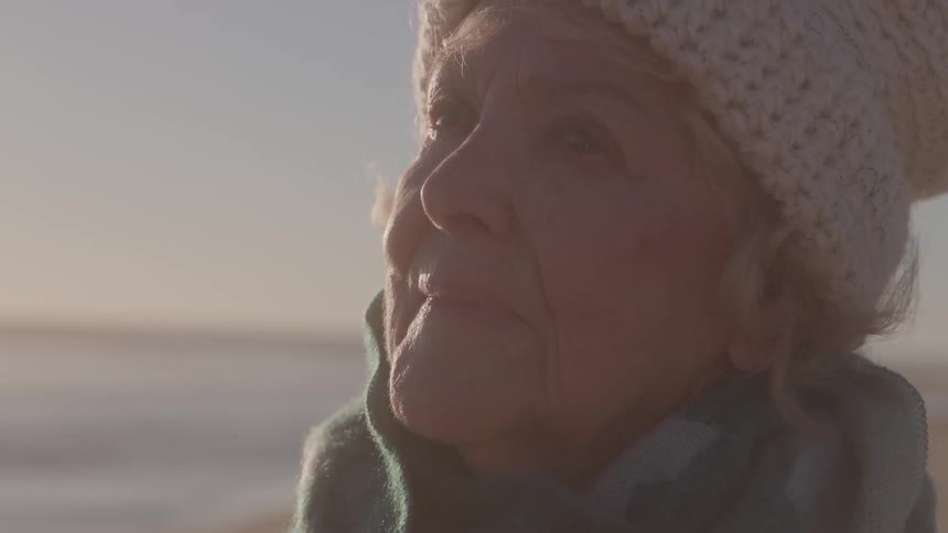 Elderly woman wearing a hat and scarf gazes at the horizon on a sunny day