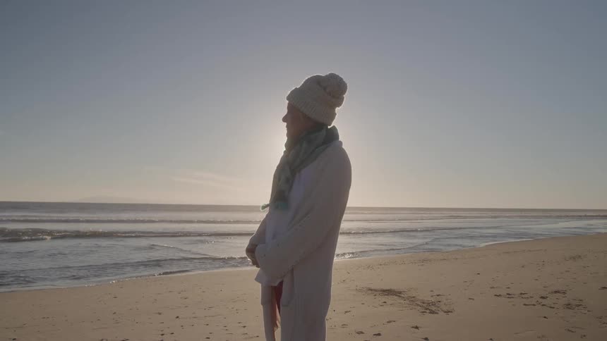 A silhouetted person wearing a knit hat and long coat stands on a wide sandy beach looking out at the ocean waves with the bright sun just above the horizon