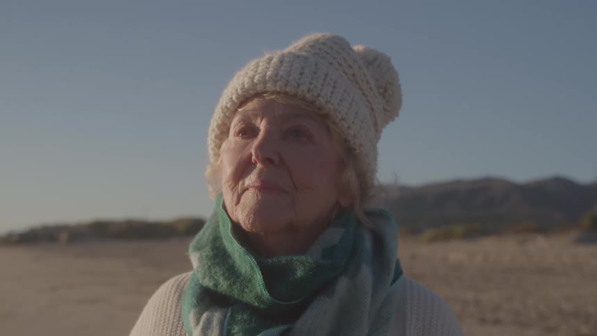 Elderly woman wearing a wool hat gazes at the horizon on a beach at sunset