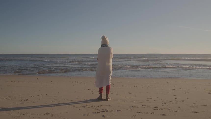 A person wearing a white knit hat red pants and a white towel wrapped around their body stands facing the ocean on a sandy beach under a clear sunny sky