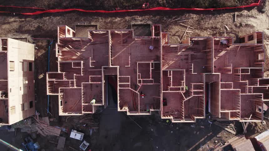 Highangle aerial view that slowly zooms out and pans upward over a construction site showing the exposed wooden framing of multiple connected residential units and surrounding earth with workers moving within the structures