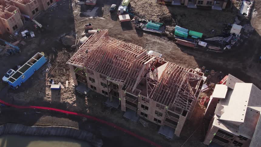 An overhead aerial view shows a multistory woodframed apartment building under construction with its roof rafters exposed surrounded by dirt and other building materials and trucks on a sunny day