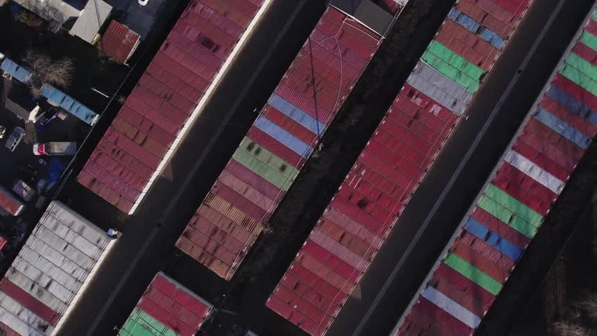 An overhead aerial view tracks over long parallel rows of shipping containers painted mostly red with some green and blue stripes separated by narrow roadways in a busy industrial yard