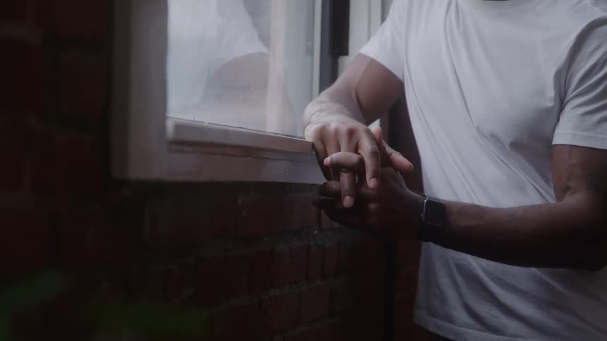 A closeup view shows a man wearing a white tshirt and a smartwatch standing indoors next to a window rubbing his hands together over a dark brick ledge