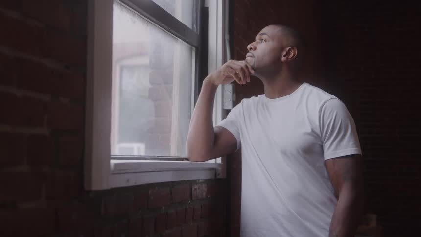 A man gazes contemplatively out a window by a dark brick wall indoors