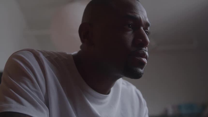 A close view shows a man with a short haircut and a beard wearing a white tshirt as he looks to the side with a serious expression in a dimly lit interior