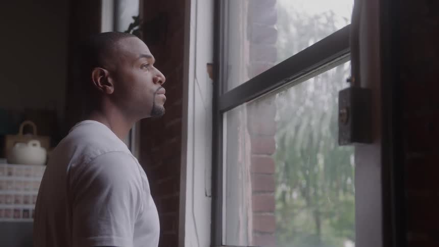 A medium close side view shows a man wearing a white tshirt looking out a large window at the rainy weather outside from a dimly lit room