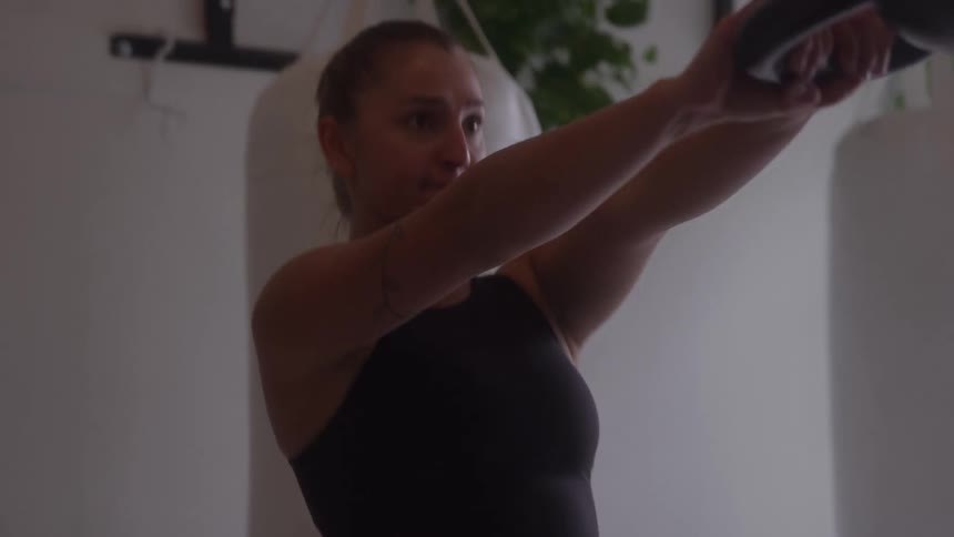 A young woman performs an exercise movement with a handheld weight inside a room with hanging heavy bags