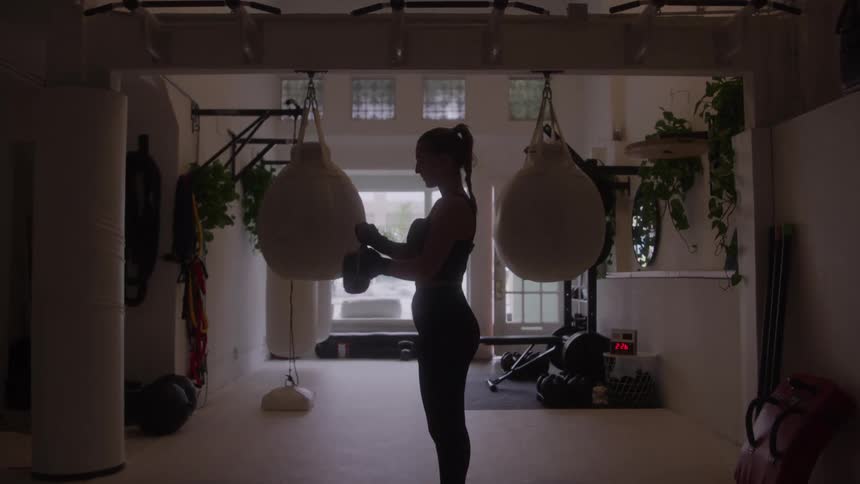 A silhouetted female boxer with a ponytail puts on her boxing gloves in a dimly lit gym standing between two large punching bags