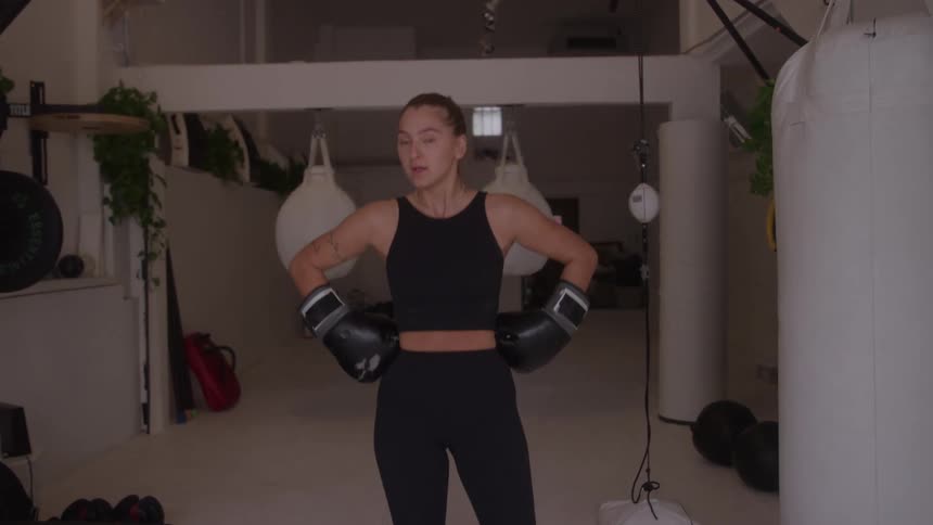 A woman in black athletic wear stands in a boxing gym with gloves on hands on hips looking focused and slightly winded