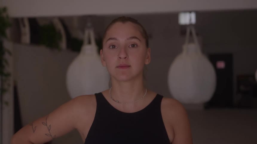 A young woman wearing a black tank top and a silver necklace looks directly at the camera and speaks in a gym with two blurred white punching bags hanging behind her