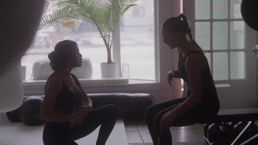 Two women in workout clothes sit and talk together inside a gym