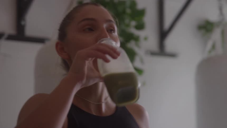 A woman in workout clothes drinks a green smoothie inside a gym