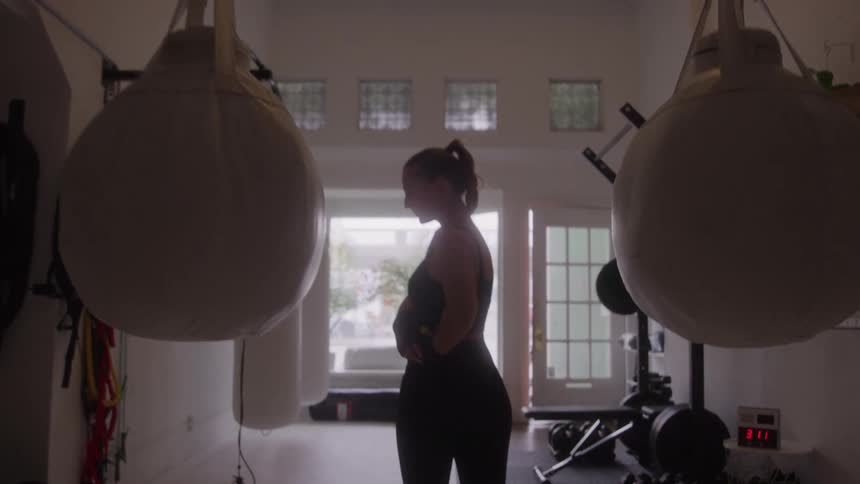 A silhouetted woman wearing black workout attire stands between two large heavy punching bags in a darkened gym adjusting her clothing with a brightly lit window and doorway in the background
