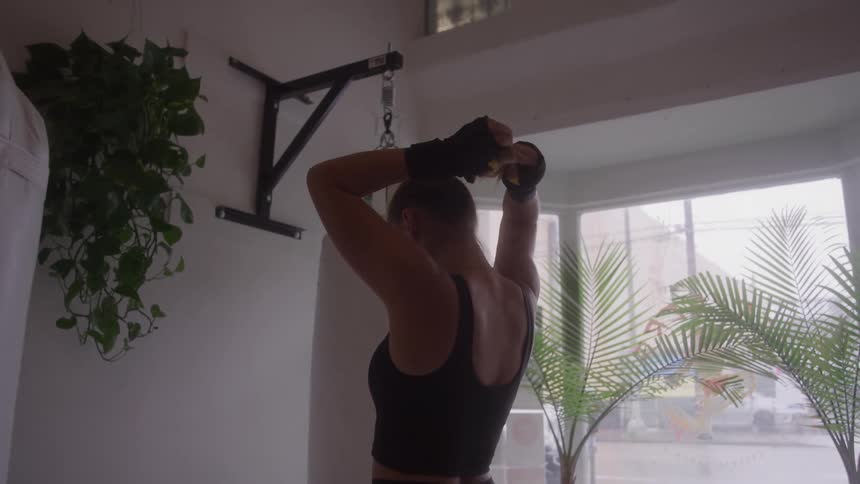A woman wearing a black tank top and boxing gloves stands with her back to the camera pulling her dark hair into a ponytail next to a white punching bag and a large window with potted palm plants in a gym