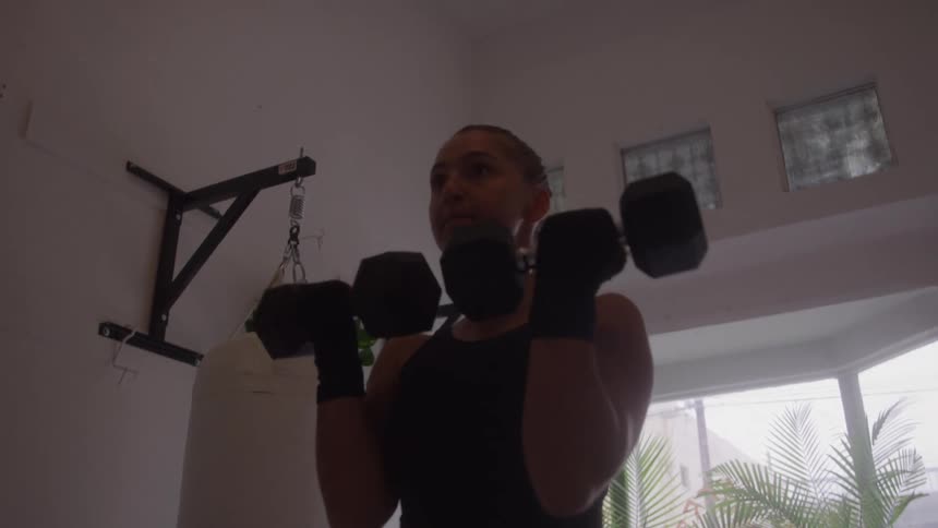 Woman lifts weights while standing near a white punching bag in a gym during the daytime