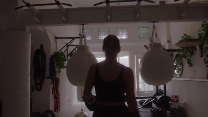 Woman wearing boxing gloves faces punching bags in a bright workout space adorned with plants