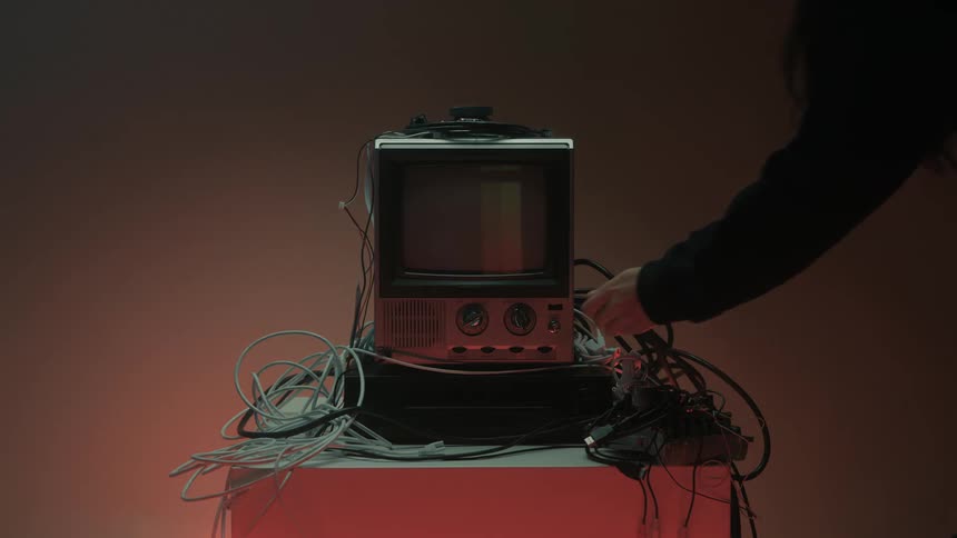 A television set displaying a color test pattern and surrounded by numerous wires with a hand reaching in to adjust a control knob on the front panel