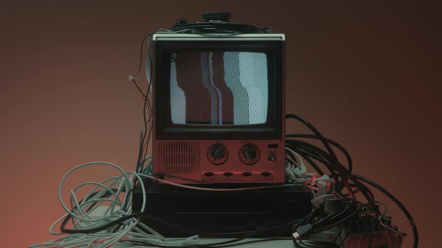 A small antique television sits on a black box surrounded by many cables and displays a vertical rolling pattern of noise and static on its screen