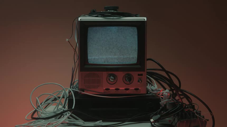A hand reaches out to turn on a vintage television set surrounded by tangled wires and the screen flickers to life with static
