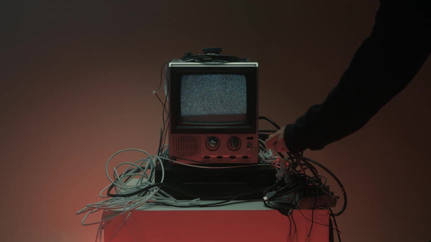 Hand adjusts a knob while turning on an old television on a table with cables