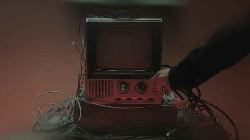 An antique television among a pile of wires displays visual noise as a human hand reaches into the frame and manipulates a button to turn it off