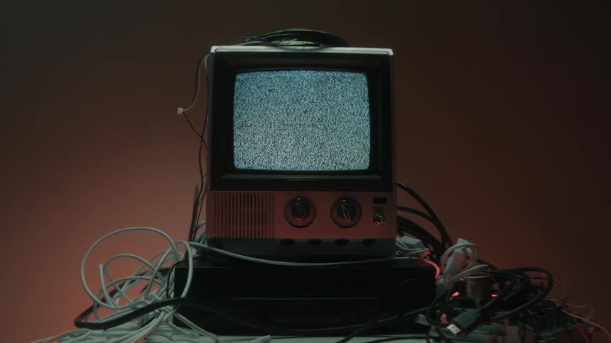 A television set displaying a screen full of moving visual noise surrounded by a tangle of cables and resting on a lower dark device on an illuminated stand