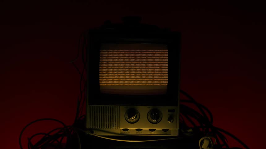 A retrostyle television set displaying continuous lines of yellow electronic characters on a dark screen sitting on a lower device and surrounded by coiled cables all within a dimly redlit space