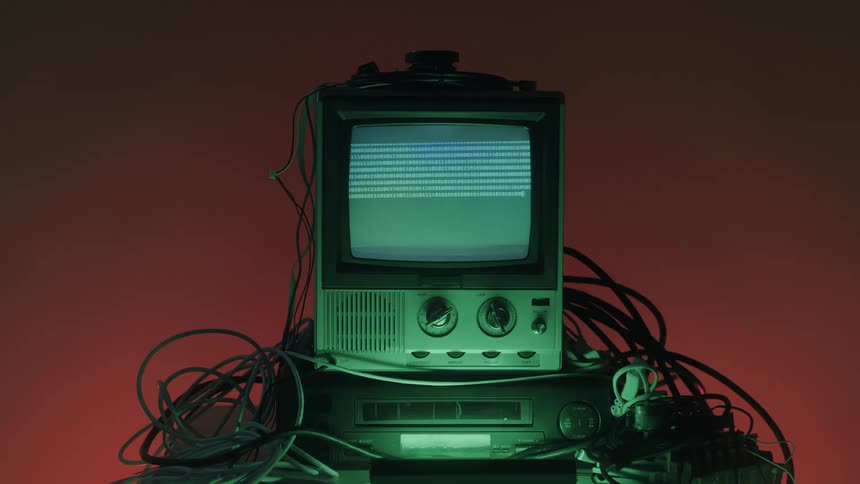 A vintage television set displaying rapidly changing digital text or electronic signal noise on a bluegreen screen urrounded by a mass of cables and lit by red and green ambient light