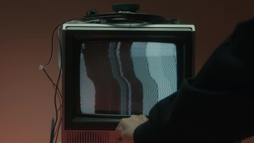 A person in a black longsleeve shirt turns on an old tv inside a room and finetunes it until rainbow colors appear on the screen