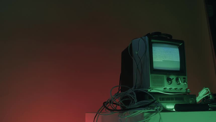 A vintage television set surrounded by a mess of wires displays lines of code in a room with dramatic red and green lighting