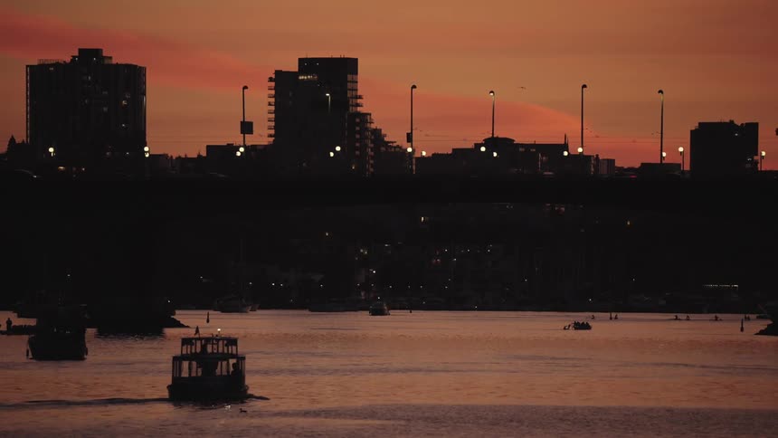 Boats sailing under a bridge at sunset with buildings in the background