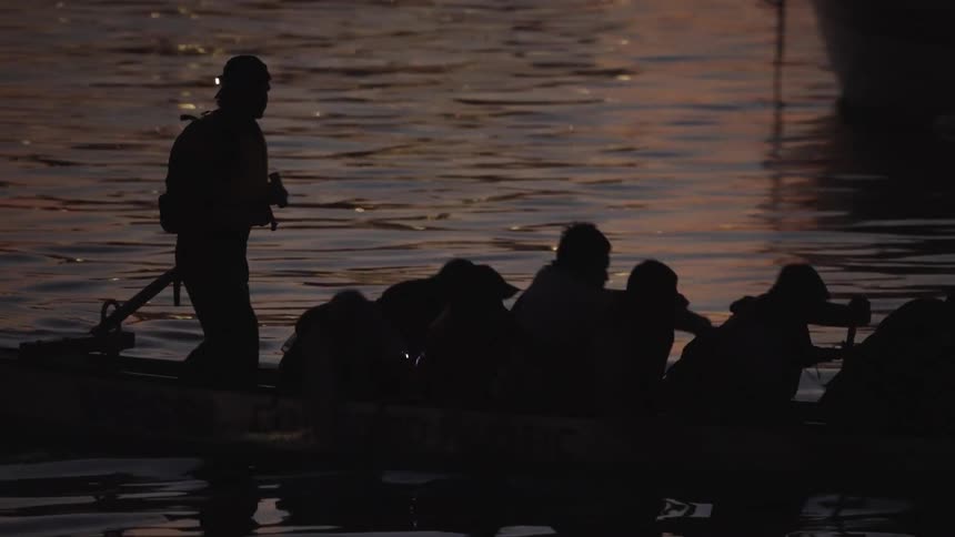 This lowlight shot shows the silhouette of a dragon boat on the water at night with a group of people seated and paddlinga single standing figure in the stern who appears to be wearing a headlamp steers or commands the vessel while