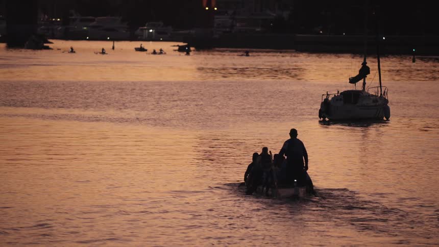 This lowlight distant shot captures a small dragon boat with a crew paddling away from the camera in a body of water silhouetted against a deep orange or amber sunsettwilight glowin the middleground a sailboat is anchored or moored a