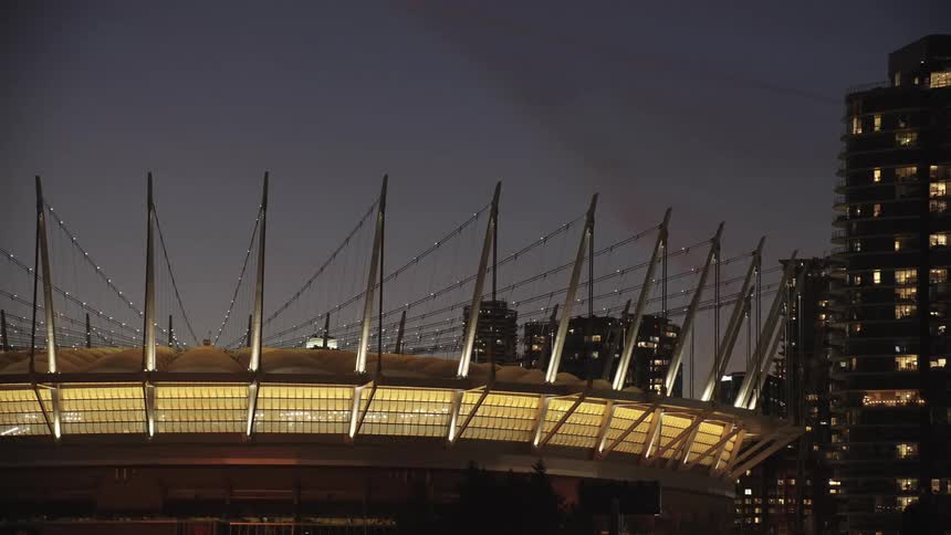 The video shows a modern stadium at night its distinctive cablesupported roof structure brightly illuminated with a warm golden light it is set against a dark night sky and flanked by the tall dark silhouettes of city highrise buildings with window