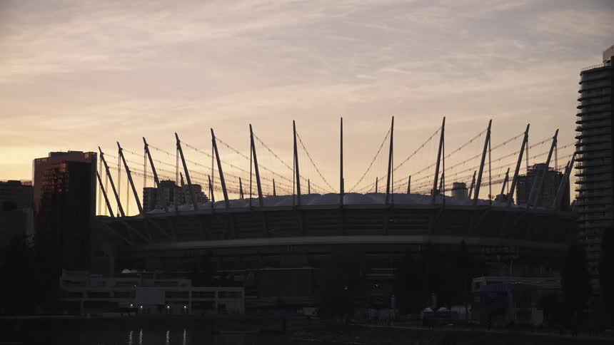 This static wide shot captures the silhouette of a large modern stadium with a prominent tensile roof structure at dusk or sunrisethe stadium is framed by tall residential buildings on either side and reflects faintly on the body of water