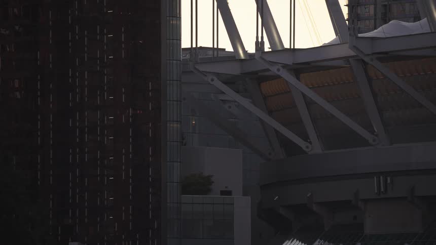 This shot features the juxtaposition of three distinct modern urban structures under a bright skyon the left is the dark gridded facade of a skyscraper while the right features the massive exposed steel framework of a stadium or large civ